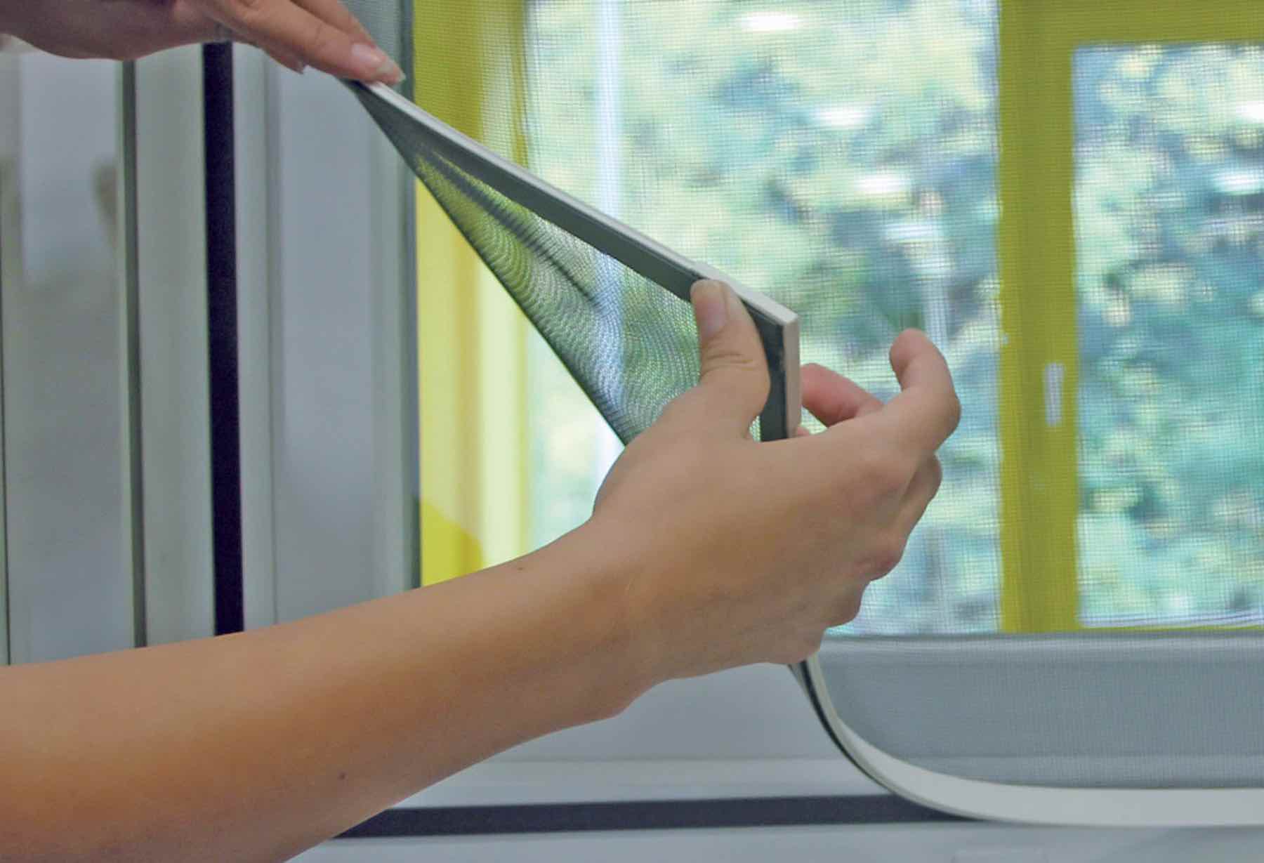 Two hands lifting a magnetic window screen away from the magnetic strip on a window frame
